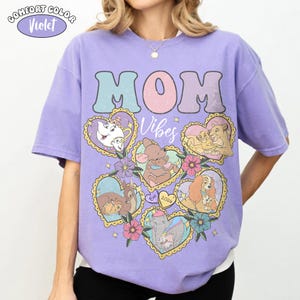 Pu&ograve; includere: Una maglietta Comfort Colors viola con la scritta "MOM" in colori pastello. La maglietta presenta illustrazioni a forma di cuore di personaggi Disney e le parole "Mom Vibes".