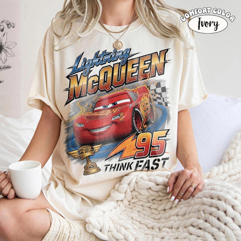 May include: Ivory-colored t-shirt featuring a graphic of Lightning McQueen from the Cars movie. The design includes the text "Lightning McQueen", "95", and "Think Fast". The shirt also has a Comfort Color label.