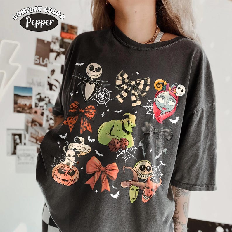 May include: A charcoal gray short-sleeve t-shirt featuring a graphic print of characters from The Nightmare Before Christmas, including Jack Skellington, Sally, and Oogie Boogie, interspersed with various bows, spiderwebs, and bats.