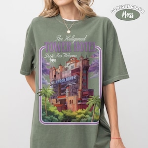 Op de afbeelding: Olijfgroen T-shirt met een afbeelding van het Hollywood Tower Hotel, met de tekst "The Hollywood Tower Hotel Drop-Ins Welcome 1994". Het ontwerp is omlijst door een paarse rand, met palmbomen en een bewolkte lucht.