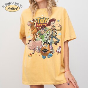 May include: Mustard-colored t-shirt featuring a graphic of characters from the animated film "Toy Story." The design includes Woody, Buzz Lightyear, and other characters. The words "TOY STORY" are printed above the characters. The shirt is a Comfort Colors brand.