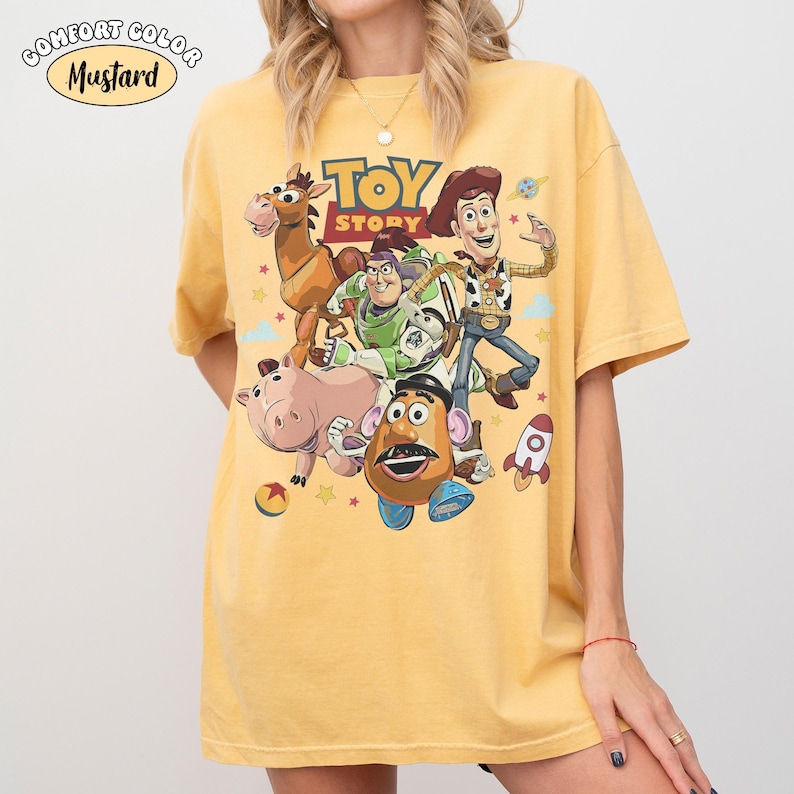 May include: Mustard-colored t-shirt featuring a graphic of characters from the animated film "Toy Story." The design includes Woody, Buzz Lightyear, and other characters. The words "TOY STORY" are printed above the characters. The shirt is a Comfort Colors brand.
