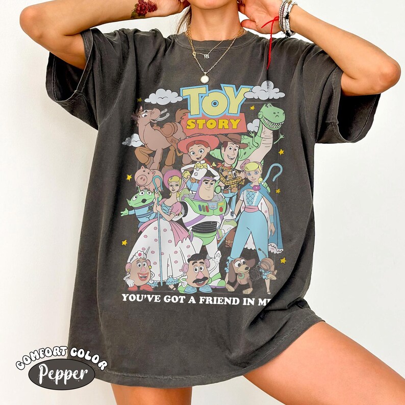 K&ouml;nnte beinhalten: Ein anthrazitfarbenes Kurzarm-T-Shirt mit einem bunten Grafikdruck der Toy Story-Figuren, darunter Woody, Buzz Lightyear und Bo Peep. Der Text lautet TOY STORY und YOU'VE GOT A FRIEND IN ME. Das T-Shirt hat eine lockere, entspannte Passform.