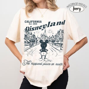 May include: Ivory-colored t-shirt with a Disneyland graphic. The design features a street scene with Mickey Mouse and the text "CALIFORNIA Disneyland Est. 1955" and "The happiest place on earth."
