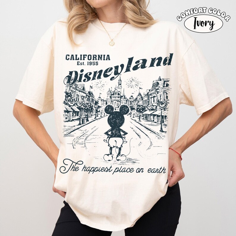 May include: Ivory-colored t-shirt with a Disneyland graphic. The design features a street scene with Mickey Mouse and the text "CALIFORNIA Disneyland Est. 1955" and "The happiest place on earth."