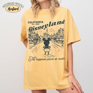 May include: Mustard-colored t-shirt with a Disneyland graphic. The graphic features a street scene with Mickey Mouse and the text "California Disneyland Est. 1955" and "The happiest place on earth."
