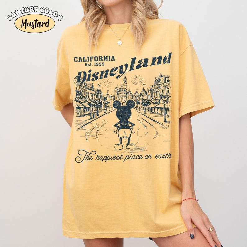 May include: Mustard-colored t-shirt with a Disneyland graphic. The graphic features a street scene with Mickey Mouse and the text "California Disneyland Est. 1955" and "The happiest place on earth."