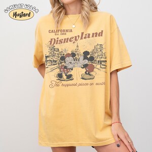 May include: A mustard yellow short-sleeve t-shirt featuring a graphic print of Mickey and Minnie Mouse. The text reads California Est. 1955 Disneyland The happiest place on earth. The design includes a line drawing of a theme park street scene.