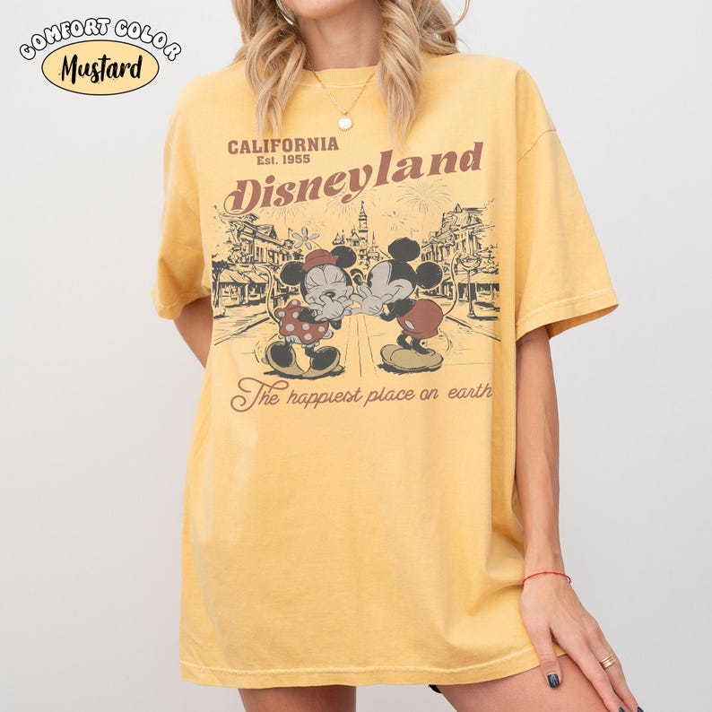 May include: A mustard yellow short-sleeve t-shirt featuring a graphic print of Mickey and Minnie Mouse. The text reads California Est. 1955 Disneyland The happiest place on earth. The design includes a line drawing of a theme park street scene.