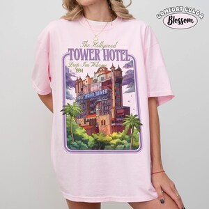 Op de afbeelding: Een lichtroze Comfort Colors t-shirt met een afbeelding van het Hollywood Tower Hotel. Het ontwerp bevat de tekst "The Hollywood Tower Hotel" en "Drop-Ins Welcome 1994". Het shirt heeft een relaxte pasvorm en korte mouwen.