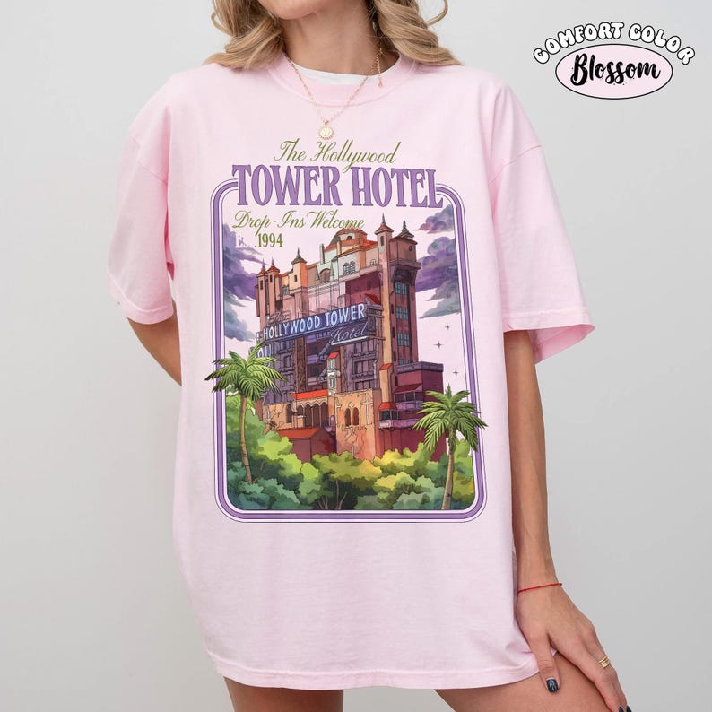Op de afbeelding: Een lichtroze Comfort Colors t-shirt met een afbeelding van het Hollywood Tower Hotel. Het ontwerp bevat de tekst "The Hollywood Tower Hotel" en "Drop-Ins Welcome 1994". Het shirt heeft een relaxte pasvorm en korte mouwen.