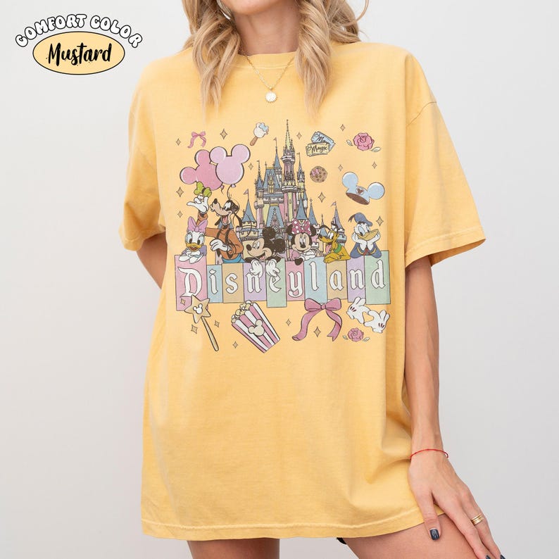 Peut inclure: T-shirt jaune moutarde avec un motif Disneyland. Le motif repr&eacute;sente un ch&acirc;teau, des personnages Disney et le mot "Disneyland" en couleurs pastel. Le t-shirt est de la marque Comfort Colors.
