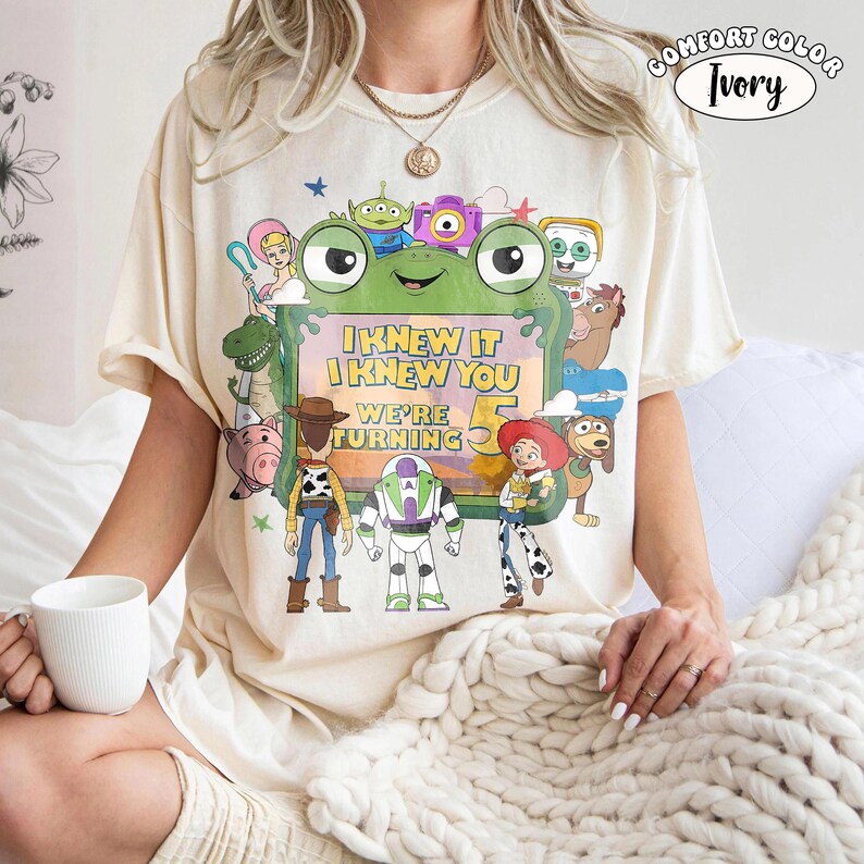 Puede incluir: Una camiseta de manga corta color marfil con un estampado gr&aacute;fico de varios personajes animados que rodean un marco verde con forma de rana. El texto dentro del marco dice I KNEW IT I KNEW YOU WE'RE TURNING 5. Se ve una etiqueta Comfort Color Ivory.