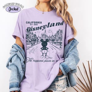 May include: Orchid Comfort Colors t-shirt featuring a Disneyland graphic. The design includes the text "CALIFORNIA Disneyland Est. 1955" above a black and white illustration of Mickey Mouse in front of a street scene. The shirt also has the text "The happiest place on er".