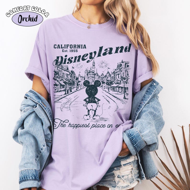 May include: Orchid Comfort Colors t-shirt featuring a Disneyland graphic. The design includes the text "CALIFORNIA Disneyland Est. 1955" above a black and white illustration of Mickey Mouse in front of a street scene. The shirt also has the text "The happiest place on er".