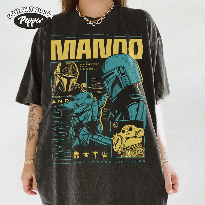 K&ouml;nnte beinhalten: Dunkelgraues Comfort Colors T-Shirt mit einem grafischen Design mit dem Text "MANDO" in Gelb. Das Design enth&auml;lt Illustrationen eines Mandalorianer-Helms und Grogu, mit dem Text "THE LEGEND CONTINUES."