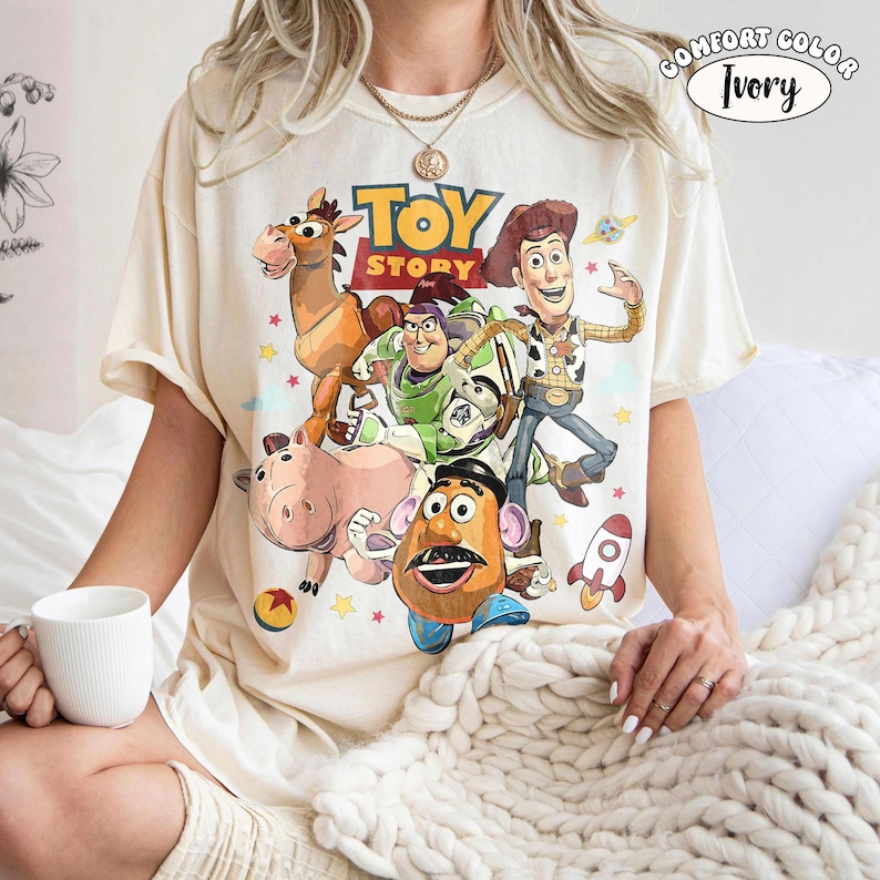 May include: Ivory-colored t-shirt featuring a colorful graphic of characters from the animated film "Toy Story." The design includes Woody, Buzz Lightyear, Mr. Potato Head, and other characters. The shirt has short sleeves and a crew neck.
