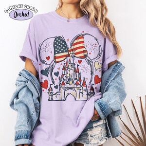 May include: A light purple t-shirt with a graphic featuring a castle, hearts, fireworks, and a bow with an American flag design. The shirt has the text "Comfort Color Orchid" in the upper left corner.