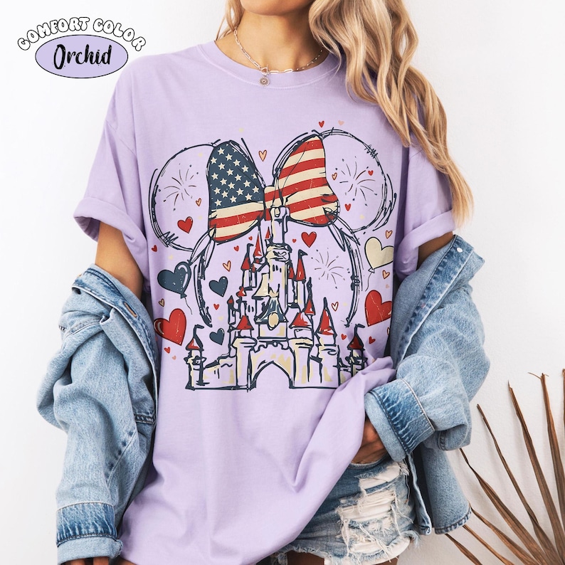 May include: A light purple t-shirt with a graphic featuring a castle, hearts, fireworks, and a bow with an American flag design. The shirt has the text "Comfort Color Orchid" in the upper left corner.