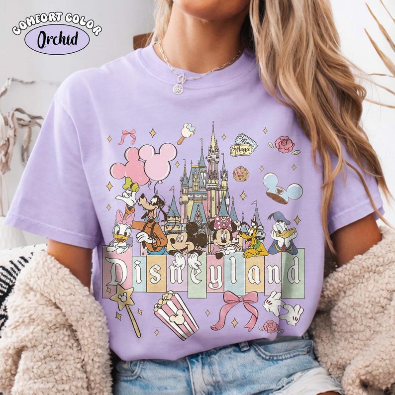 Peut inclure: T-shirt lilas Comfort Colors avec un motif Disneyland. Le design comprend des personnages de dessins anim&eacute;s, un ch&acirc;teau et le mot "Disneyland" en couleurs pastel. &Eacute;l&eacute;ments suppl&eacute;mentaires : Mickey, pop-corn et n&oelig;uds.