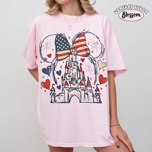 May include: A light pink t-shirt featuring a graphic of a castle, a bow with an American flag design, and Mickey Mouse ears. The design includes red, white, and blue hearts and fireworks. The text "COMFORT COLOR Blossom" is printed at the top.