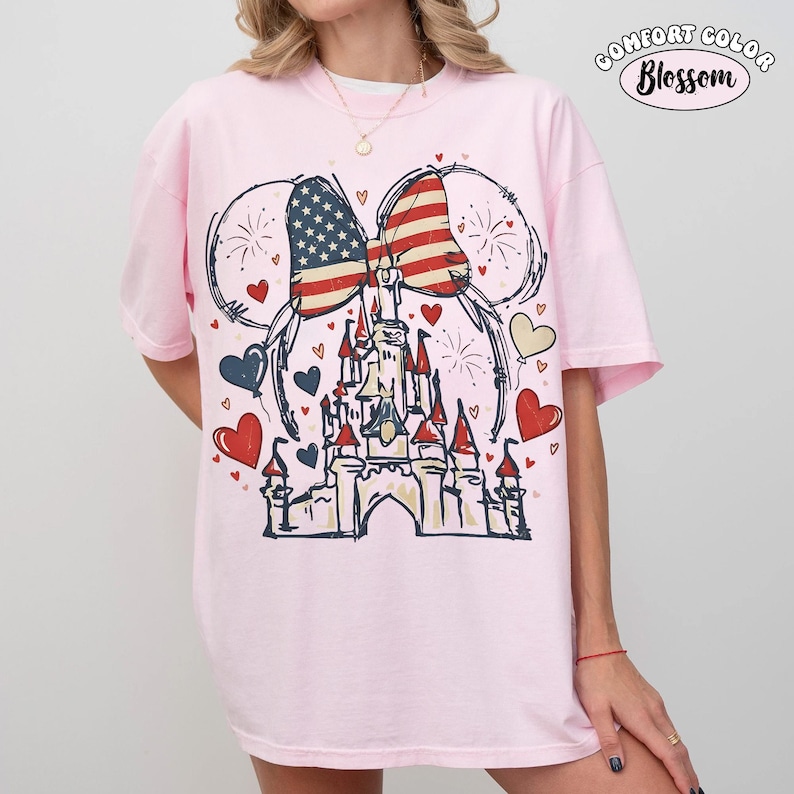 May include: A light pink t-shirt featuring a graphic of a castle, a bow with an American flag design, and Mickey Mouse ears. The design includes red, white, and blue hearts and fireworks. The text "COMFORT COLOR Blossom" is printed at the top.