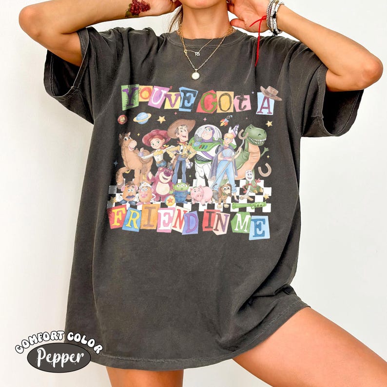 May include: Dark gray Comfort Colors t-shirt featuring a colorful graphic of Toy Story characters with the text "YOU'VE GOT A FRIEND IN ME". The shirt has a relaxed fit and a vintage look.