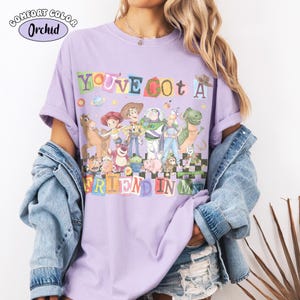 May include: A lilac Comfort Colors t-shirt featuring a colorful graphic of characters from the movie Toy Story. The text "YOU'VE GOT A FRIEND IN ME" is displayed in a playful font. The shirt has short sleeves and a relaxed fit.