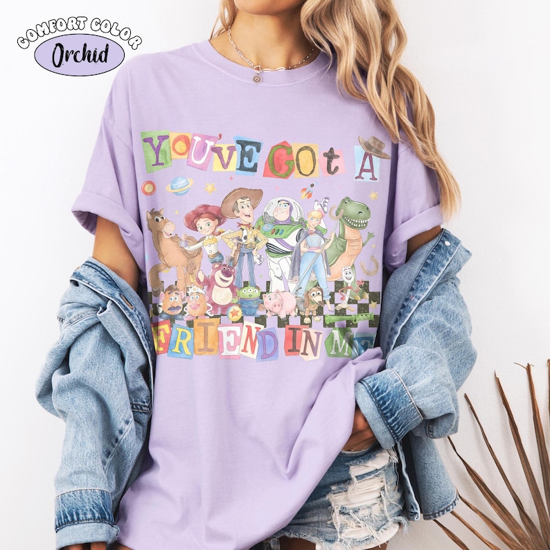 May include: A lilac Comfort Colors t-shirt featuring a colorful graphic of characters from the movie Toy Story. The text "YOU'VE GOT A FRIEND IN ME" is displayed in a playful font. The shirt has short sleeves and a relaxed fit.