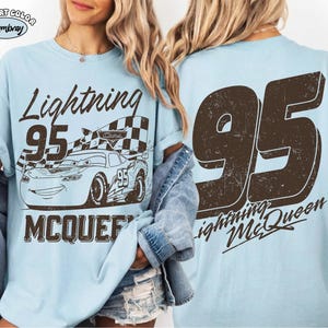 May include: Light blue t-shirt featuring a graphic of Lightning McQueen from the movie Cars. The front design includes the text "Lightning 95 McQueen" with an image of the race car. The back of the shirt displays a large "95" and "Lightning McQueen".