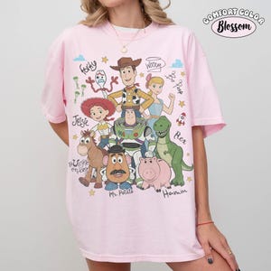 Może przedstawiać: Jasnor&oacute;żowy t-shirt z nadrukiem graficznym przedstawiającym postacie z Toy Story, w tym Chudego, Buzza Astrala, Jessie, Cienkiego, Pana Bulwę, Rexa, Hamm, Pastereczkę i Sztućka, wraz z odręcznie napisanymi imionami i tekstem Comfort Color Blossom.