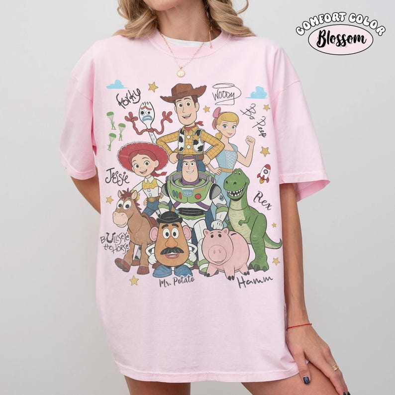 Może przedstawiać: Jasnor&oacute;żowy t-shirt z nadrukiem graficznym przedstawiającym postacie z Toy Story, w tym Chudego, Buzza Astrala, Jessie, Cienkiego, Pana Bulwę, Rexa, Hamm, Pastereczkę i Sztućka, wraz z odręcznie napisanymi imionami i tekstem Comfort Color Blossom.