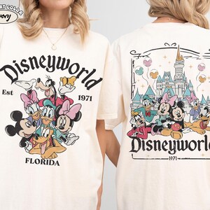 K&ouml;nnte beinhalten: Ein elfenbeinfarbenes T-Shirt mit einem Grafikdruck von Disney-Figuren wie Mickey Maus, Minnie Maus, Donald Duck, Daisy Duck, Goofy und Pluto. Die Vorderseite zeigt den Text Disneyworld Est 1971 Florida. Die R&uuml;ckseite zeigt das Schloss.