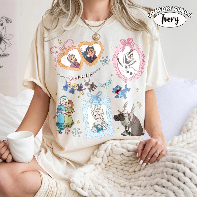 May include: Ivory-colored t-shirt featuring various characters and elements from the animated film "Frozen." The design includes framed portraits of characters, snowflakes, and the word "FROZEN." The shirt is a Comfort Colors brand.