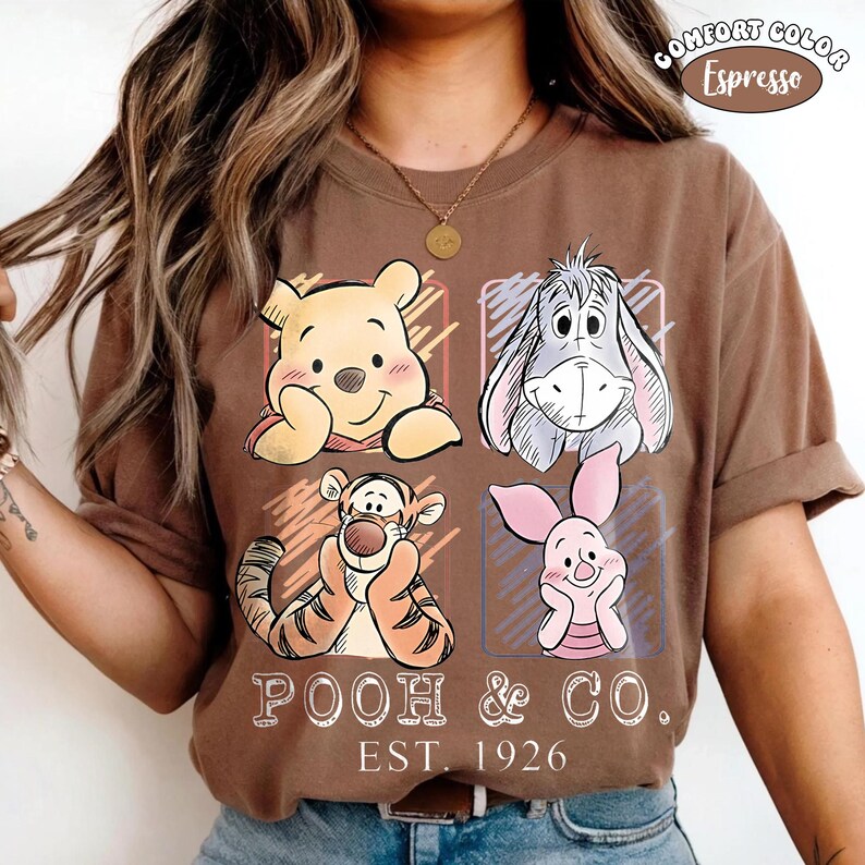 May include: Espresso-colored t-shirt featuring four cartoon characters: Winnie the Pooh, Eeyore, Tigger, and Piglet. The text "POOH & CO. EST. 1926" is printed below the characters. The shirt has a Comfort Color label.