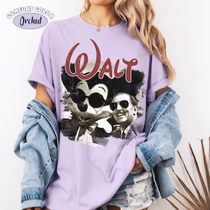 Puede incluir: Una camiseta lila Comfort Colors con un gr&aacute;fico de Mickey Mouse con gafas de sol y Walt Disney. La palabra "Walt" est&aacute; en escritura burdeos encima de la imagen. La camiseta tiene las mangas enrolladas.