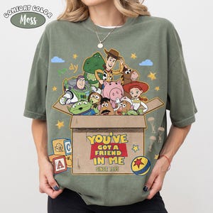 K&ouml;nnte beinhalten: Olivgr&uuml;nes Comfort Colors T-Shirt mit einem Toy Story-Motiv. Das Design zeigt Woody, Buzz Lightyear und andere Charaktere in einer Box mit dem Text "YOU'VE GOT A FRIEND IN ME SINCE 1995".