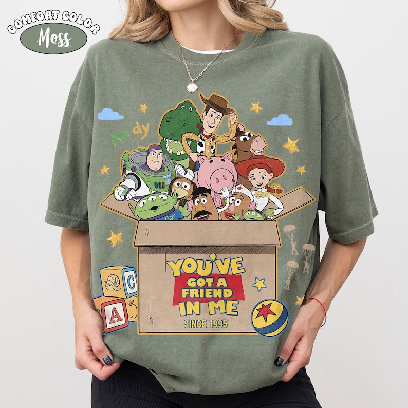 K&ouml;nnte beinhalten: Olivgr&uuml;nes Comfort Colors T-Shirt mit einem Toy Story-Motiv. Das Design zeigt Woody, Buzz Lightyear und andere Charaktere in einer Box mit dem Text "YOU'VE GOT A FRIEND IN ME SINCE 1995".