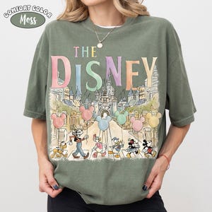 Pu&ograve; includere: T-shirt verde muschio con la scritta "THE DISNEY" in colori pastello sopra un'illustrazione di personaggi Disney e un castello. La maglietta ha una vestibilit&agrave; ampia e maniche corte.