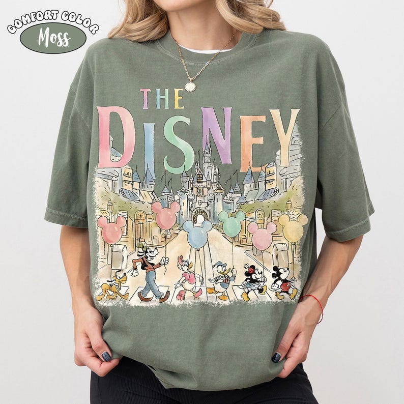 Pu&ograve; includere: T-shirt verde muschio con la scritta "THE DISNEY" in colori pastello sopra un'illustrazione di personaggi Disney e un castello. La maglietta ha una vestibilit&agrave; ampia e maniche corte.