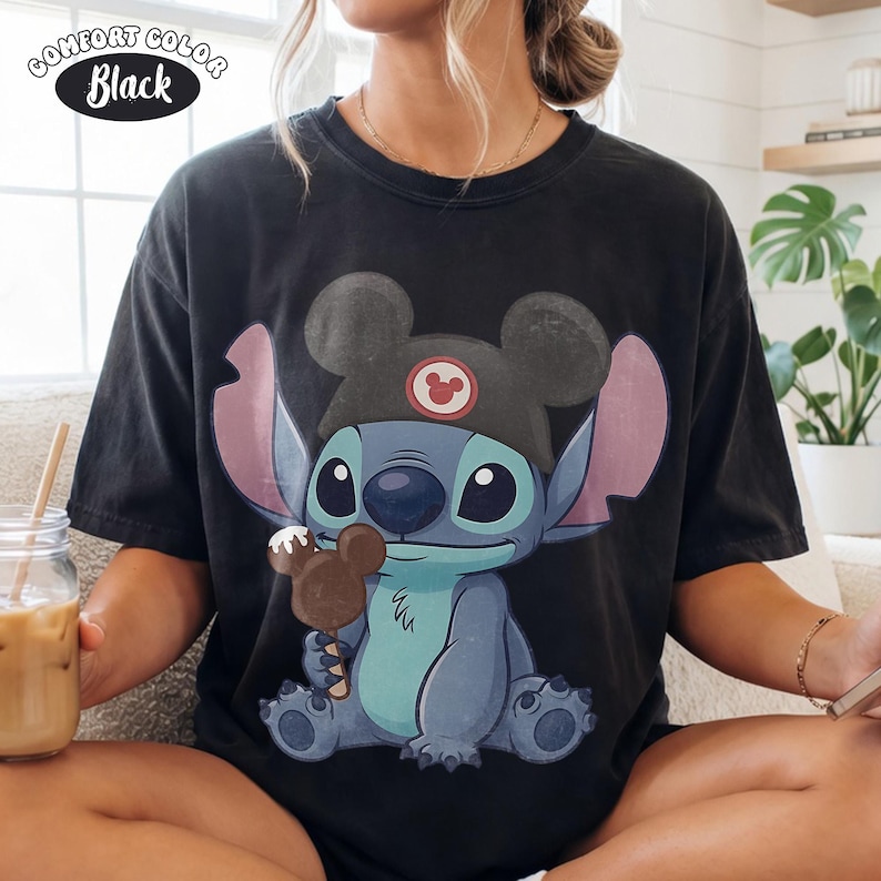 May include: A black short-sleeve t-shirt featuring a graphic of the character Stitch wearing a black mouse-ear hat and holding a chocolate-covered mouse-shaped ice cream bar. The text COMFORT COLOR Black is printed in the upper left corner.