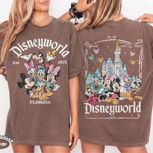 K&ouml;nnte beinhalten: Ein braunes Kurzarm-T-Shirt mit einem Grafikdruck von Disney-Figuren. Die Vorderseite zeigt Mickey, Minnie, Donald, Daisy, Goofy und Pluto mit dem Text Disneyworld Est 1971 Florida. Die R&uuml;ckseite zeigt das Schloss und die Figuren.