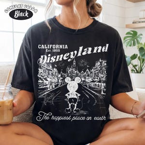 May include: Black Comfort Colors t-shirt featuring a Disneyland graphic. The design includes the text "California Est. 1955 Disneyland" with a Mickey Mouse illustration and the phrase "The happiest place on earth."