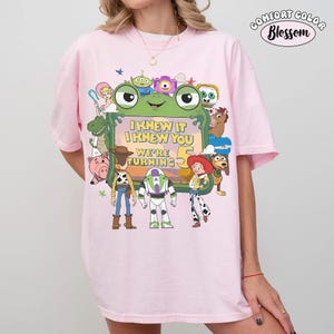 Puede incluir: Una camiseta rosa claro con un gran estampado gr&aacute;fico de varios personajes animados rodeando un marco verde con forma de rana. El texto dentro del marco dice I KNEW IT I KNEW YOU WE'RE TURNING 5. La esquina superior derecha muestra el texto COMFORT COLOR Blossom.