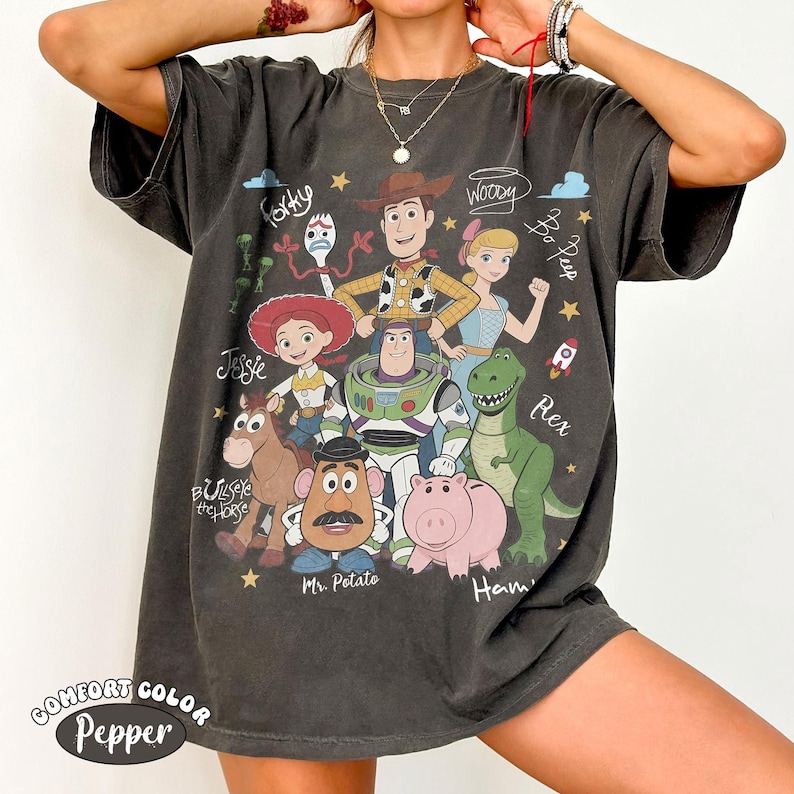 Może przedstawiać: Ciemnoszary t-shirt z kr&oacute;tkim rękawem z nadrukiem graficznym przedstawiającym postacie z Toy Story, w tym Chudego, Buzza Astrala, Jessie, Celka, Pana Bulwę, Hamm, Pastereczkę i Sztućka, z ich imionami zapisanymi kursywą.
