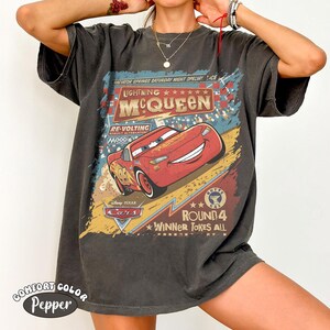 May include: Dark grey t-shirt featuring Lightning McQueen from Disney Pixar's Cars. The graphic includes the text "Lightning McQueen" and "Round 4 Winner Takes All" with a vintage, distressed design. The shirt is labeled "Comfort Color Pepper".
