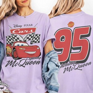 Może przedstawiać: Liliowa koszulka z Lightning McQueen z filmu Auta Disneya Pixara. Grafika z przodu zawiera logo Auta, szachownice i Lightning McQueen. Tył przedstawia dużą czerwoną liczbę 95 z imieniem Lightning McQueen.