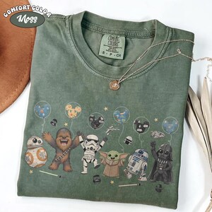May include: Green Comfort Colors t-shirt featuring Star Wars characters holding balloons. The characters include BB-8, Chewbacca, a Stormtrooper, Grogu, R2-D2, and Darth Vader. The shirt has the words "Comfort Color Moss" printed on it.