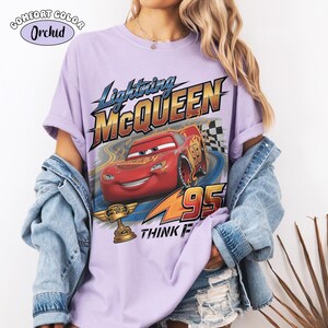 May include: A lilac Comfort Colors t-shirt featuring Lightning McQueen from the movie Cars. The graphic includes the text "Lightning McQueen" and "95", along with a racing scene and the words "THINK FAST".