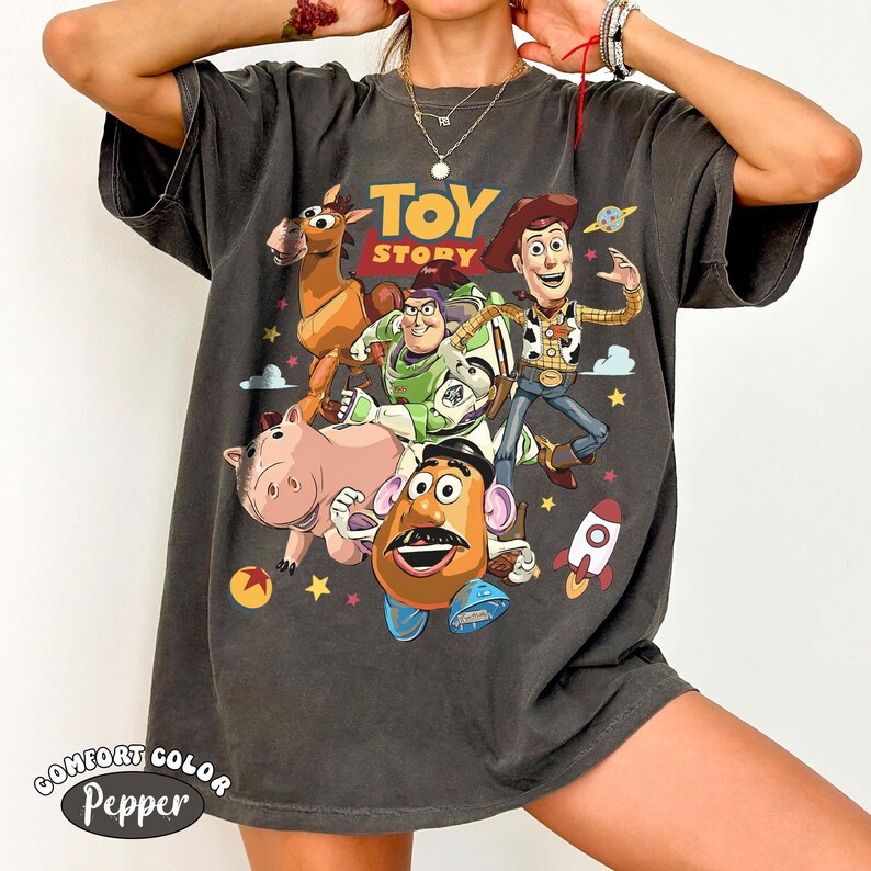 May include: Dark gray t-shirt featuring a colorful graphic of characters from the animated film "Toy Story." The design includes Woody, Buzz Lightyear, Mr. Potato Head, Hamm, and Bullseye. The shirt also has the text "TOY STORY" in yellow.